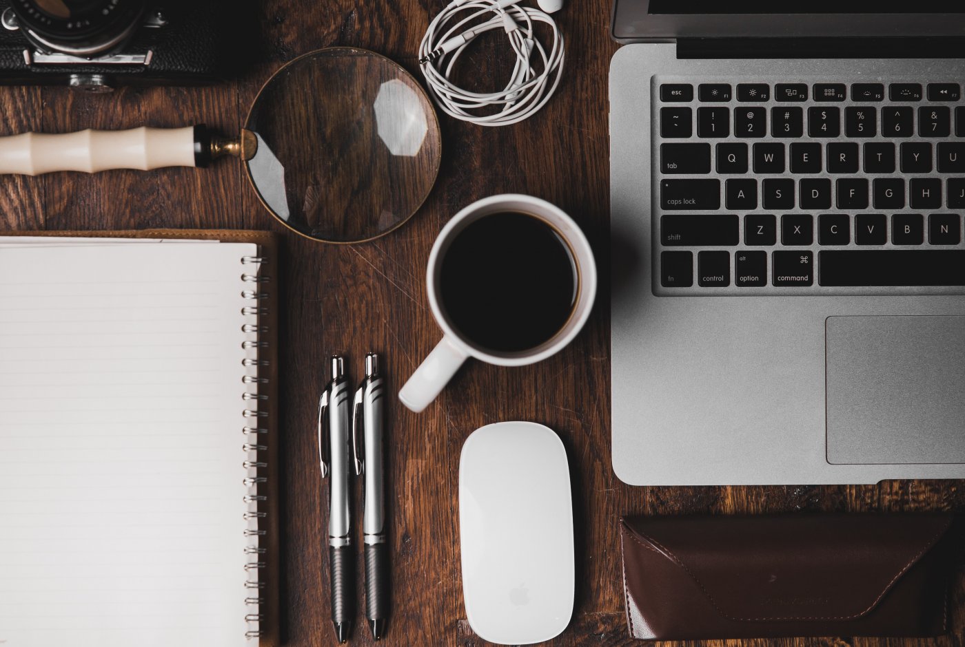 a desktop with a Mac, a cup of coffee, pens, a notepad and a magnifying glass